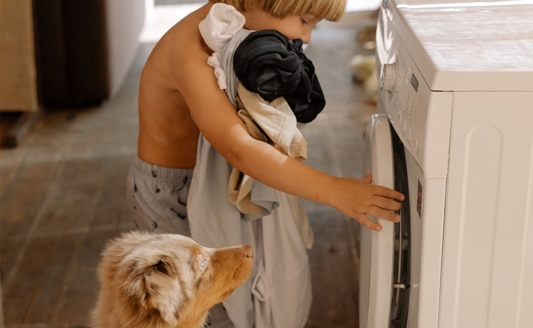 Ein Junge mit Schmutzwäsche im Arm öffnet die Waschmaschine, dabei schaut ihm ein Hund zu.